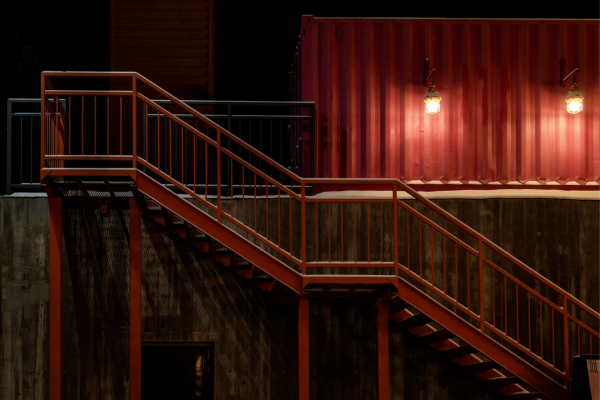 Escadaria metálica em aço corten enferrujado com corrimão laranja-ferrugem, parede de concreto bruto e container reaproveitado ao fundo iluminado por luminárias industriais, em fachada de arquitetura pós-industrial noturna