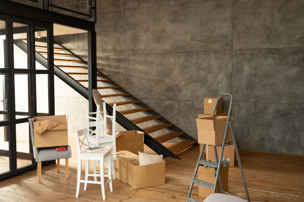 Interior de loft com parede de concreto aparente texturizado, escada metálica preta, piso de madeira natural e mezanino, durante processo de mudança com caixas empilhadas