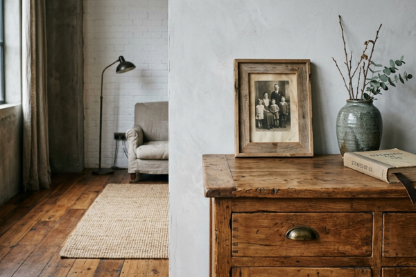 Imagem decorativa que ilustra o conceito de design afetivo sustentável aplicado a um interior contemporâneo. O canto de sala apresenta um aparador de madeira maciça restaurado com marcas de uso visíveis nas arestas. Sobre o aparador, há uma fotografia familiar emoldurada em madeira natural, um vaso de cerâmica artesanal em tom cinza-esverdeado e um livro com marcador de couro. O piso é de madeira de demolição com variações de tonalidade entre as tábuas. Um tapete de sisal trançado em tom natural cobre parte do piso. A iluminação lateral destaca a textura da madeira do aparador.