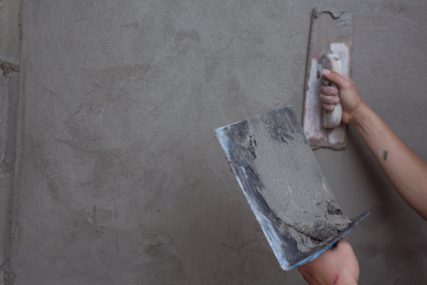 Mãos aplicando reboco artesanal com desempenadeira em parede de concreto, criando textura irregular e rugosa característica do acabamento manual